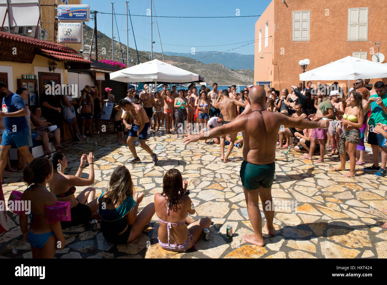 Tänzer und Publikum in Badekleidung auf gepflastertem Dorfplatz vor rötlichen Haus und Meer, Insel Kastellorizo, Dodekanes, Griechenland Stock Photo