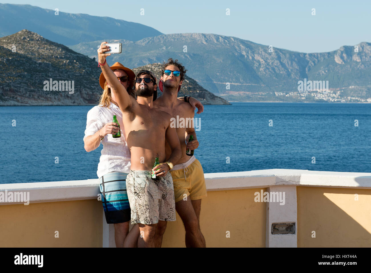 drei junge Männer in Badehosen mit Flaschen machen ein Selfie mit Meer und Küste im Hintergrund, Insel Kastellorizo, Dodekanes, Griechenland Stock Photo