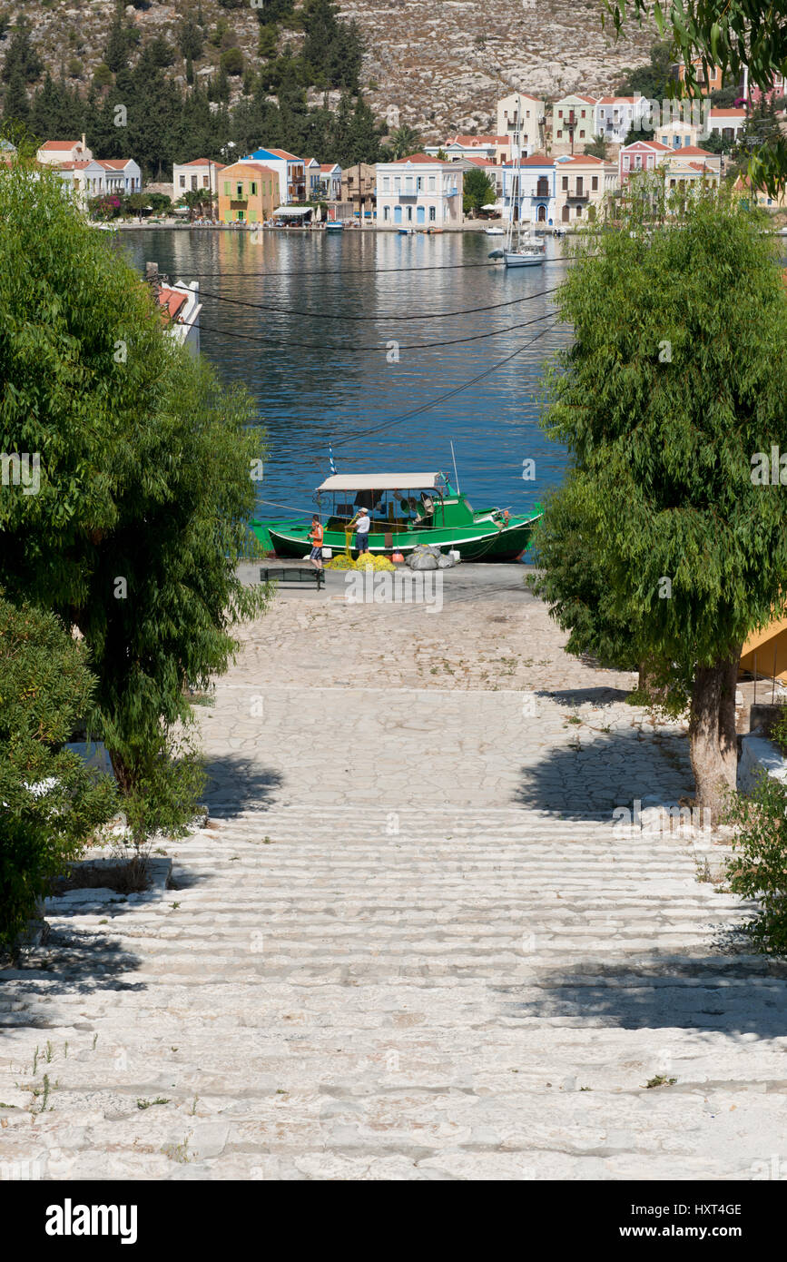 abwärts führende Treppe von Bäumen gesäumt, dahinter Hafen mit bunten Häusern und grünem Fischerboot, Insel Kastellorizo, Dodekanes, Griechenland Stock Photo