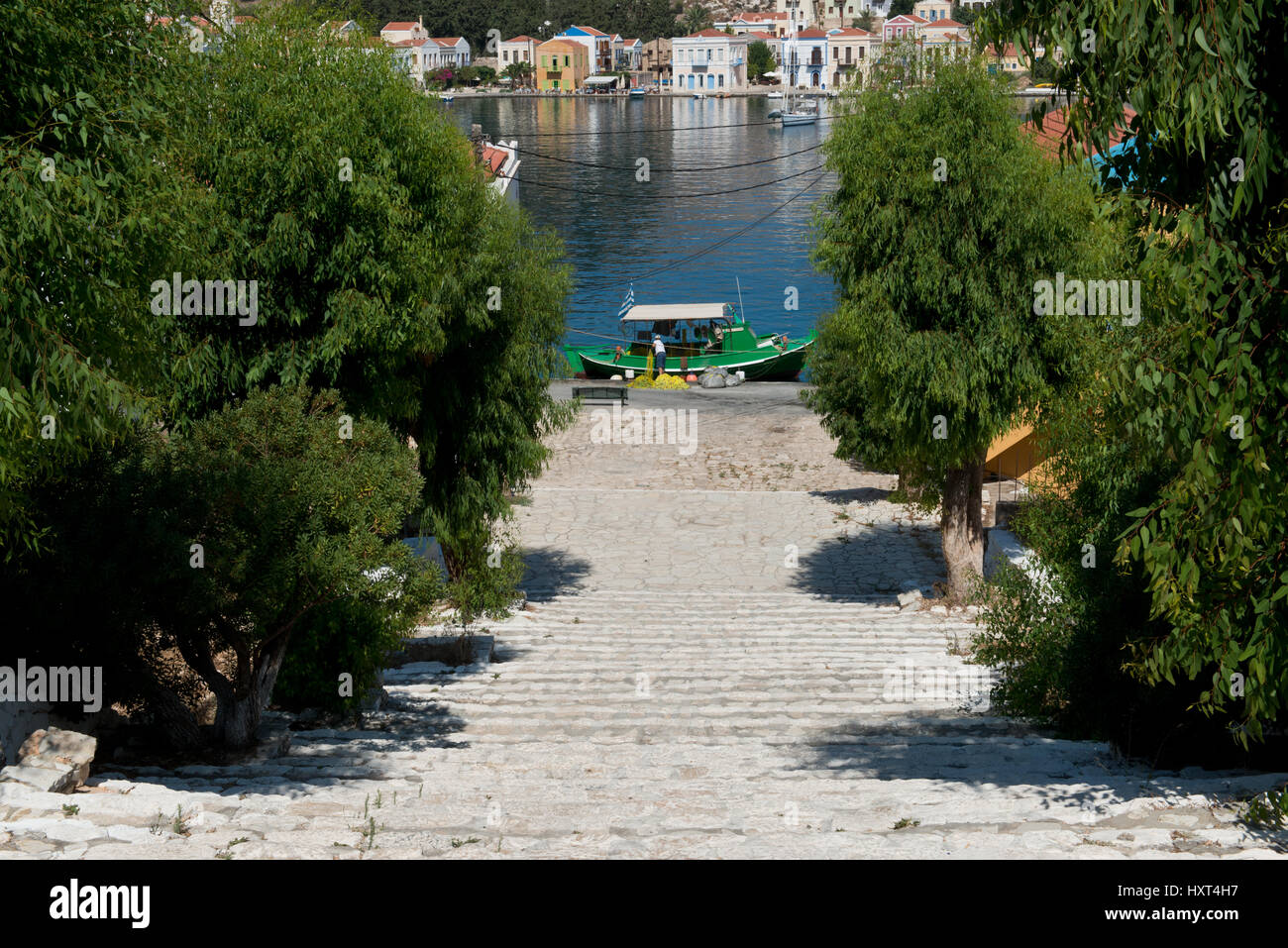 abwärts führende Treppe von Bäumen gesäumt, dahinter Hafen mit bunten Häusern und grünem Fischerboot, Insel Kastellorizo, Dodekanes, Griechenland Stock Photo