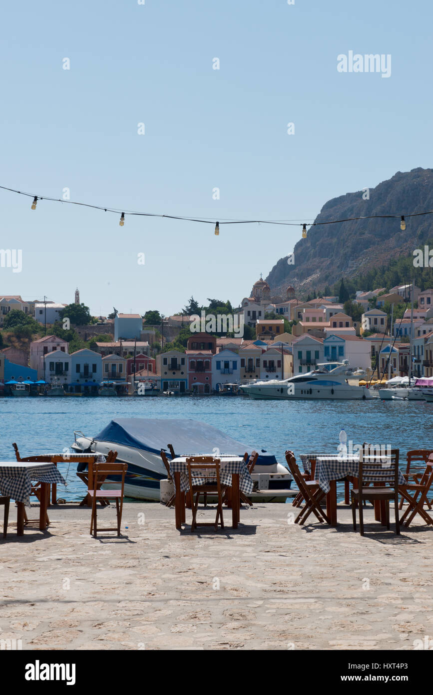 Tische und Stühle an Hafenpier, dahinter Boot, Wasser und bunte Häuser, Berge im Hintergrund, Insel Kastellorizo, Dodekanes, Griechenland Stock Photo