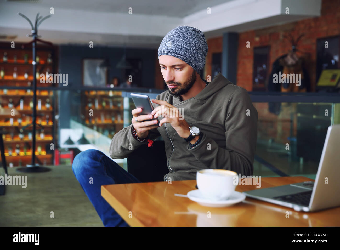 Young handsome hipster man with beard sitting in cafe talking mobile phone, holding cup of coffee and smiling. Laptop on wooden table. Stock Photo