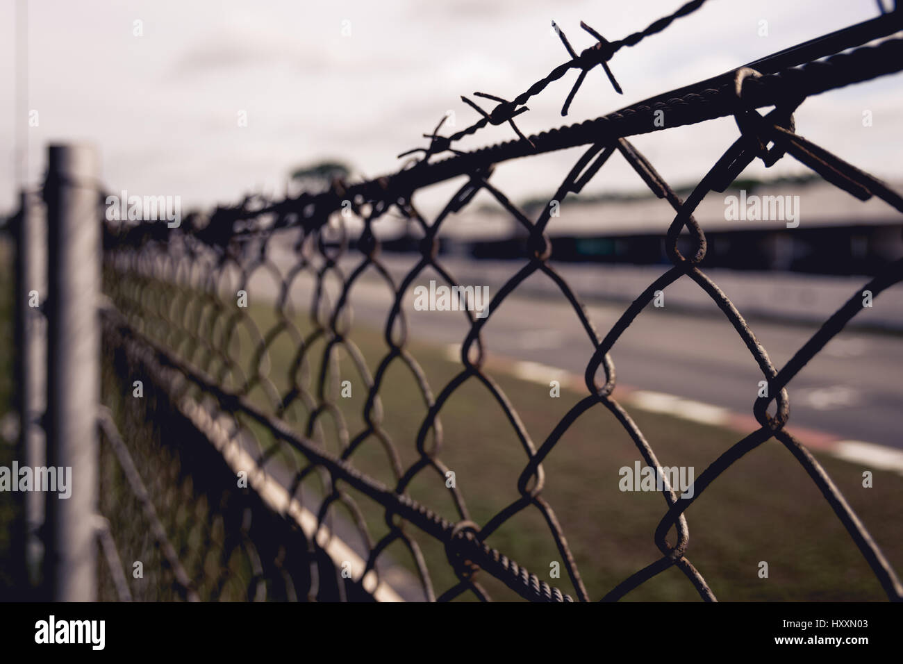 Chain-linked fence with barbed wire at top with grass and road in the background Stock Photo