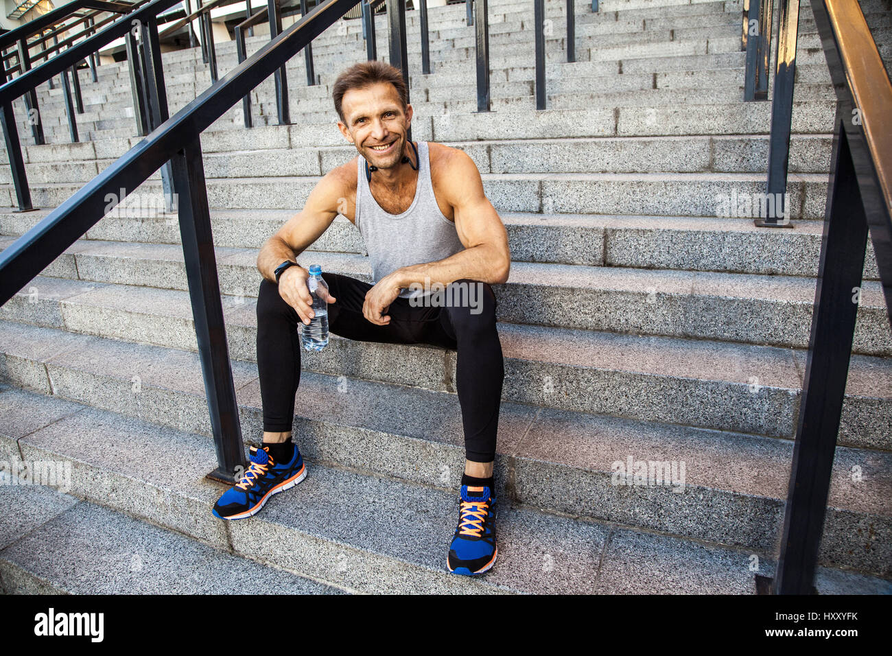 Happy smily sportsman resting and holding bottle of water, sitting on steps looking at camera and smiling . fitness, sport, exercising and people heal Stock Photo