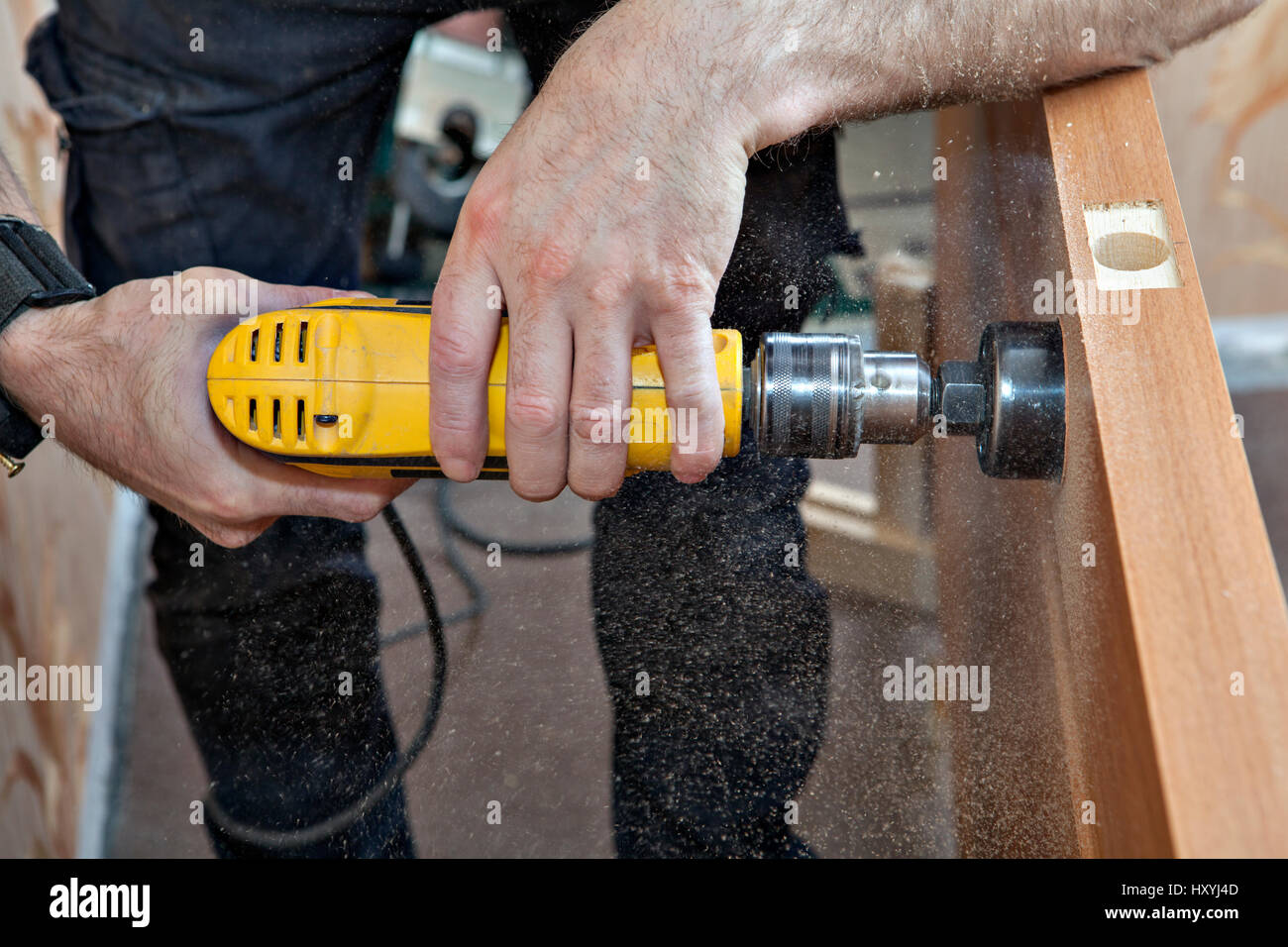 Hands woodworker with yellow electric drill, boring large hole of door lock, using hole saw