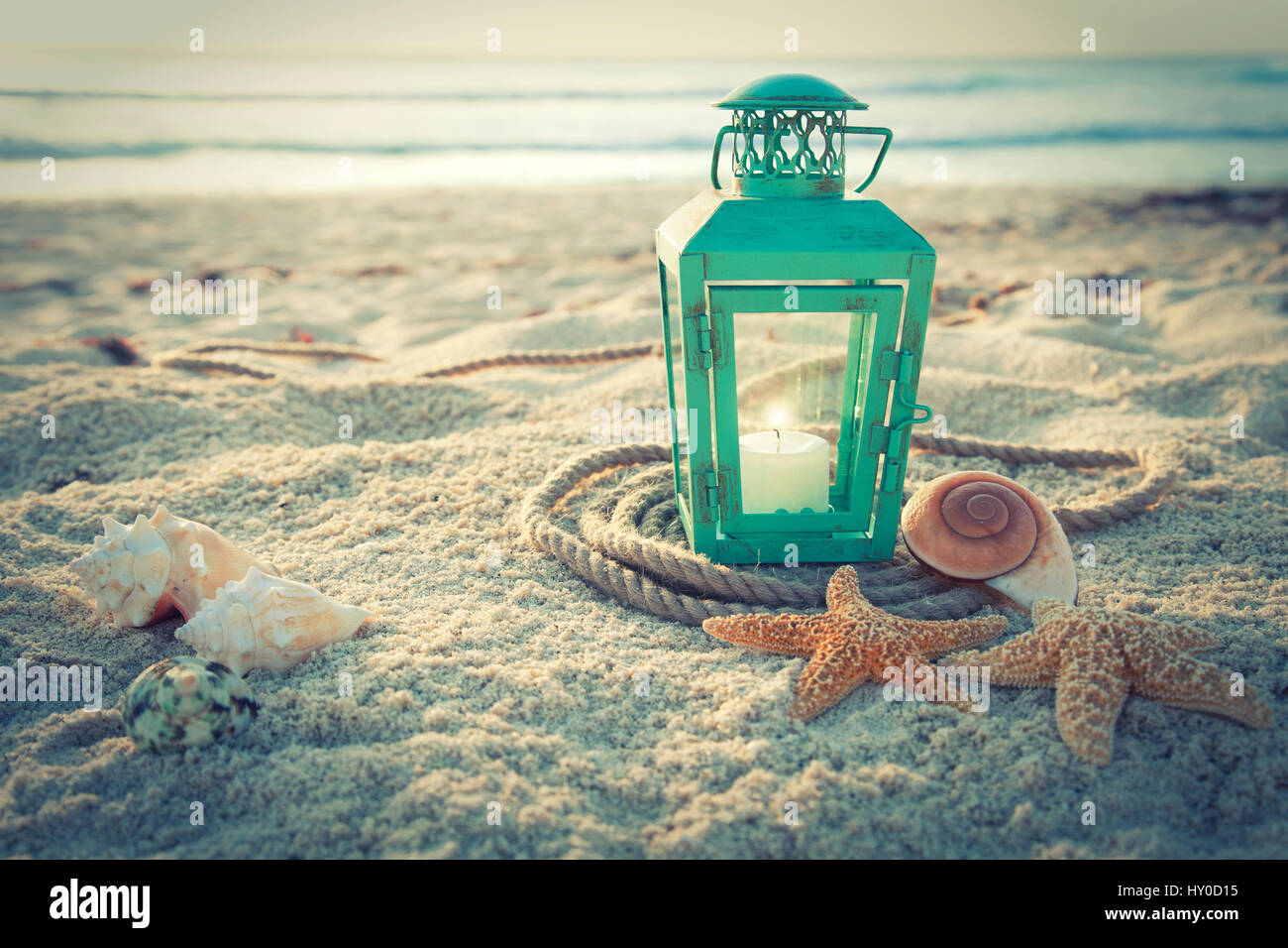 Cross-processed lantern on the beach with shells and rope at sunrise Stock Photo