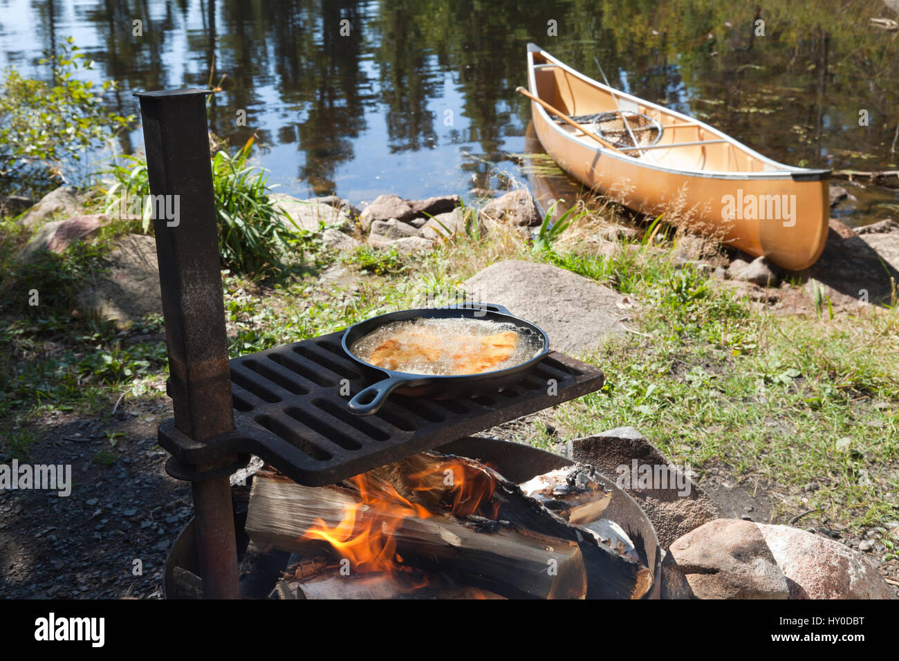 Fish cooking in a frying pan over an open fire with a canoe and northern Minnesota lake in the background Stock Photo