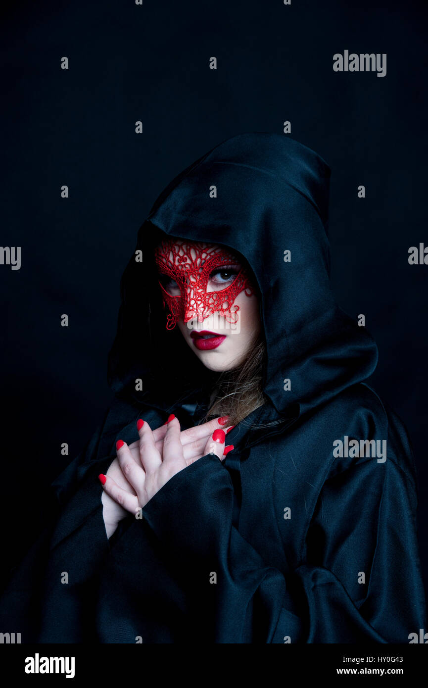 Young woman with long fair hair, wearing a red lace mask and a black, hooded cape Stock Photo