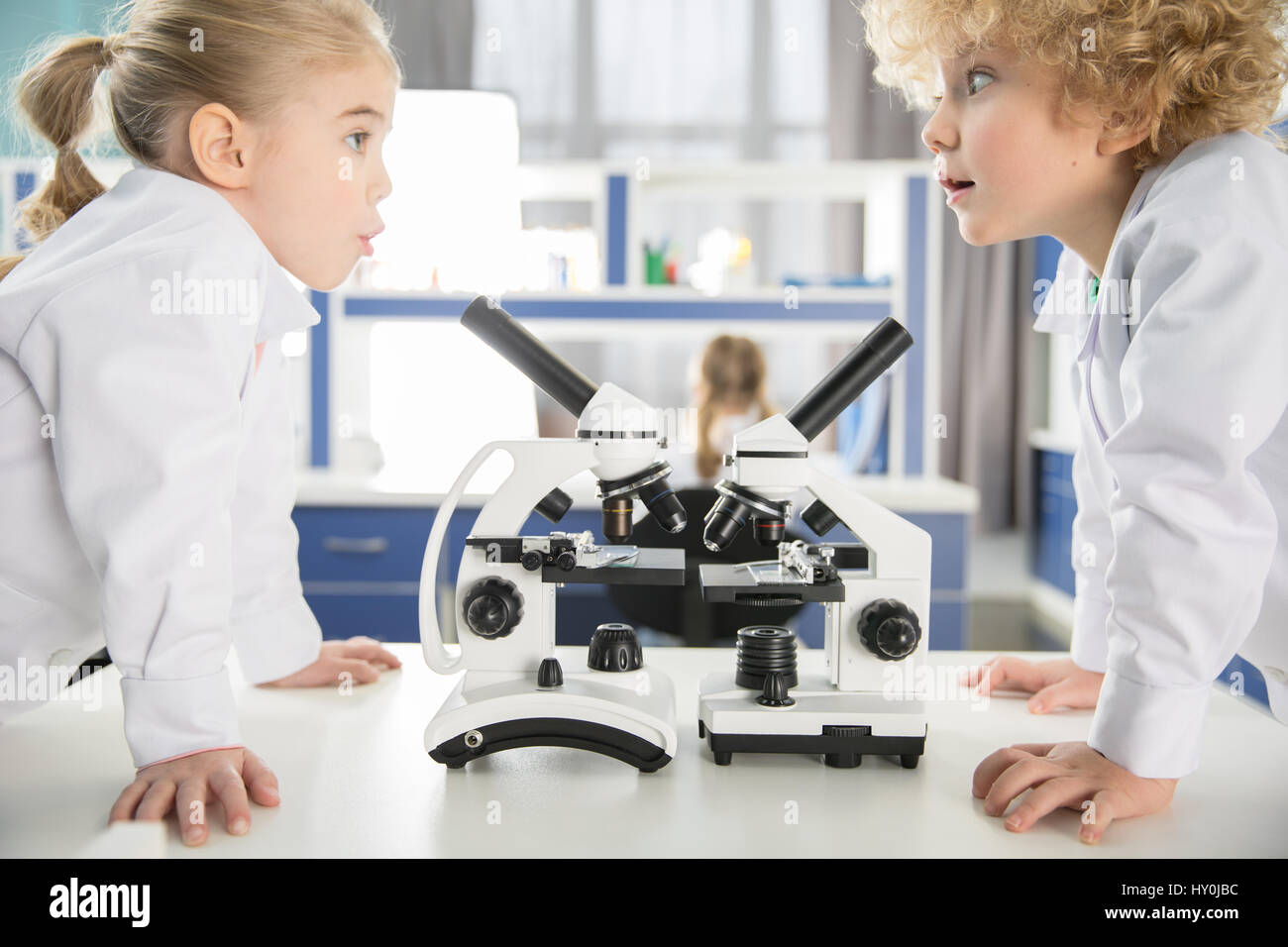 Schoolchildren in lab coats using microscopes and looking at each other in laboratory Stock Photo
