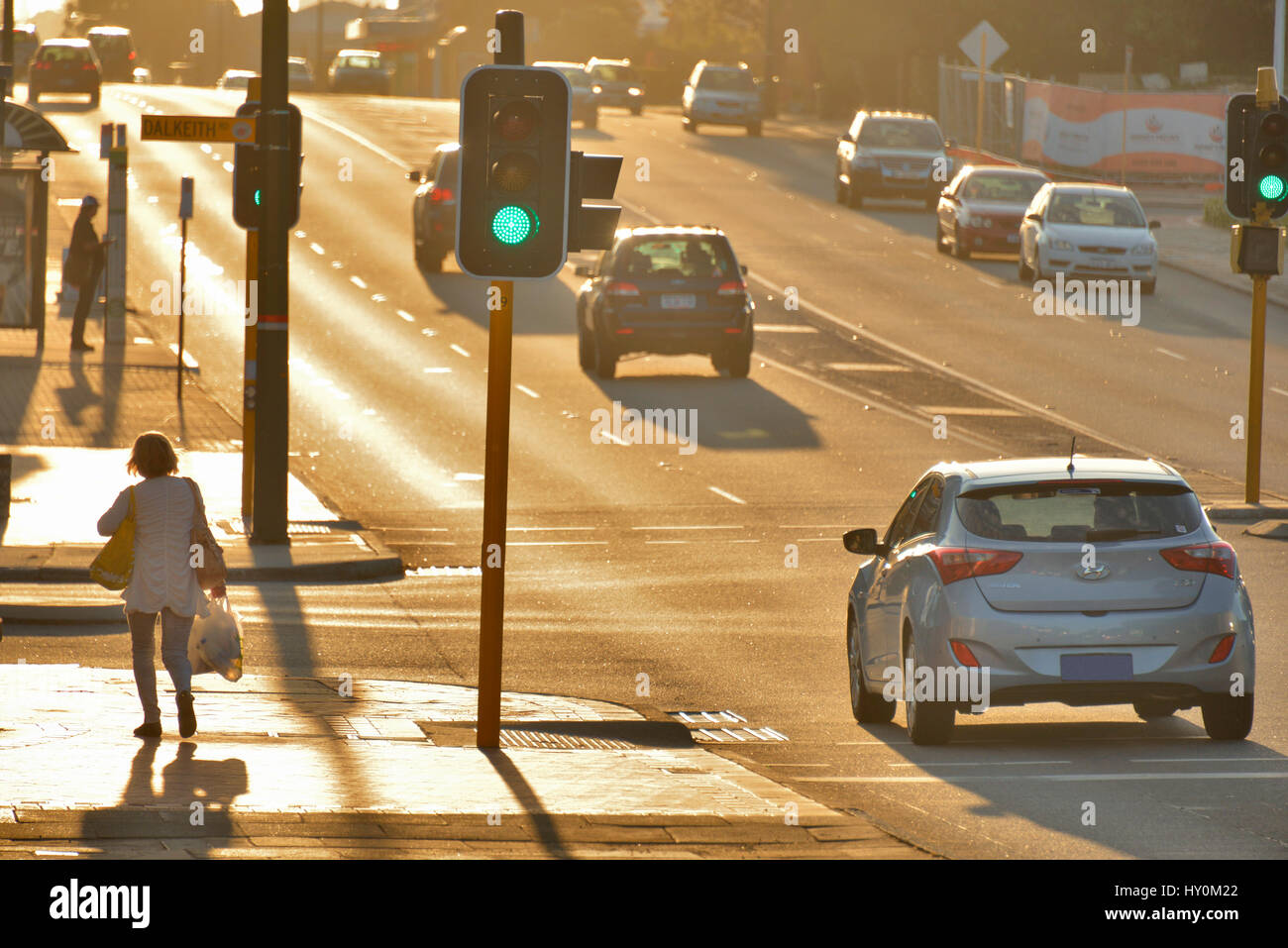 Woman carrying shopping on footpath alongside a busy road with traffic, at sunset. Stock Photo