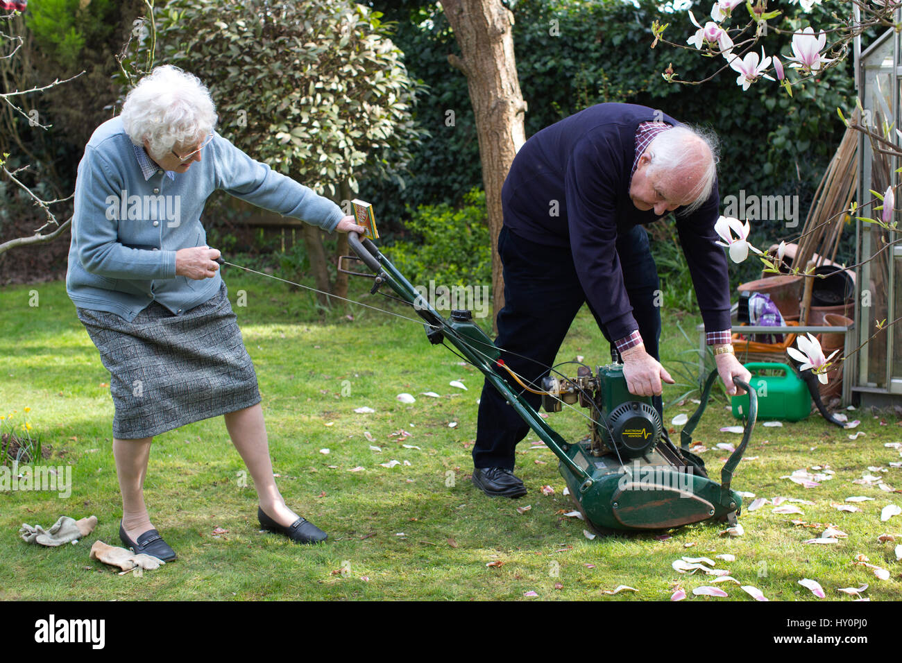 Elderly pensioners trying to start a petrol lawn mower in a English residential garden on a Spring afternoon, Surrey, England, United Kingdom Stock Photo