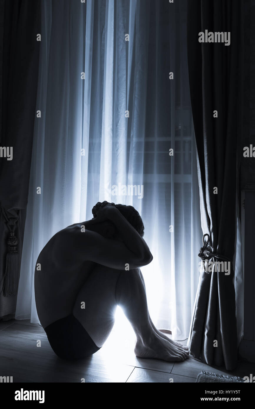 Young sad man sitting in dark room near the blue window at night Stock