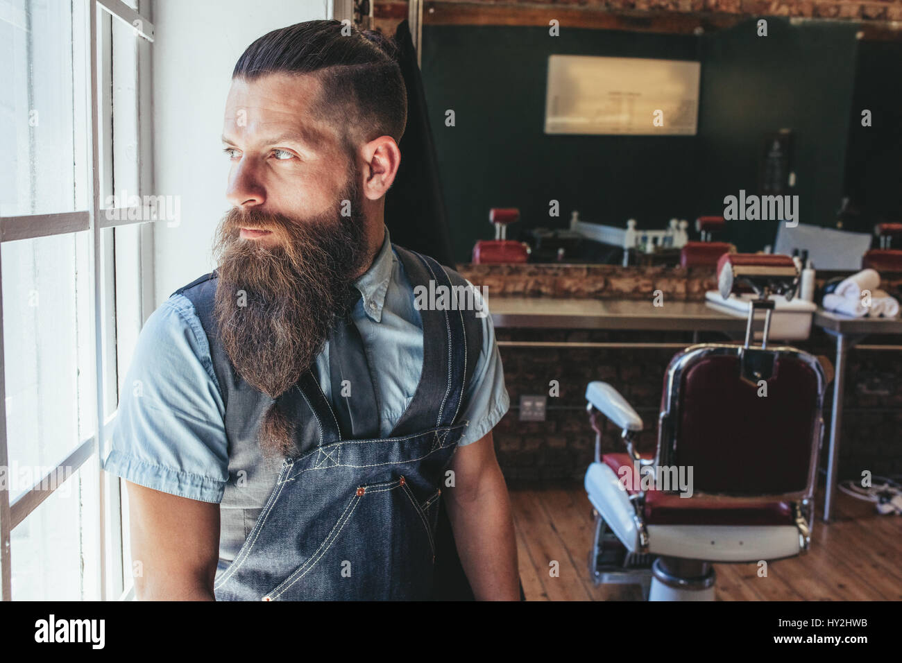 Portrait of barber with apron standing by window and looking away. Male hairdresser looking outside window and thinking. Stock Photo