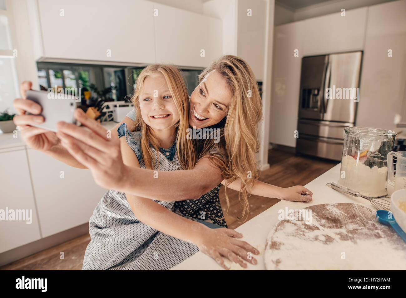 Cute little girl and her beautiful mother taking selfie and smiling preparing the dough for baking in the kitchen. Happy young family of mother and da Stock Photo