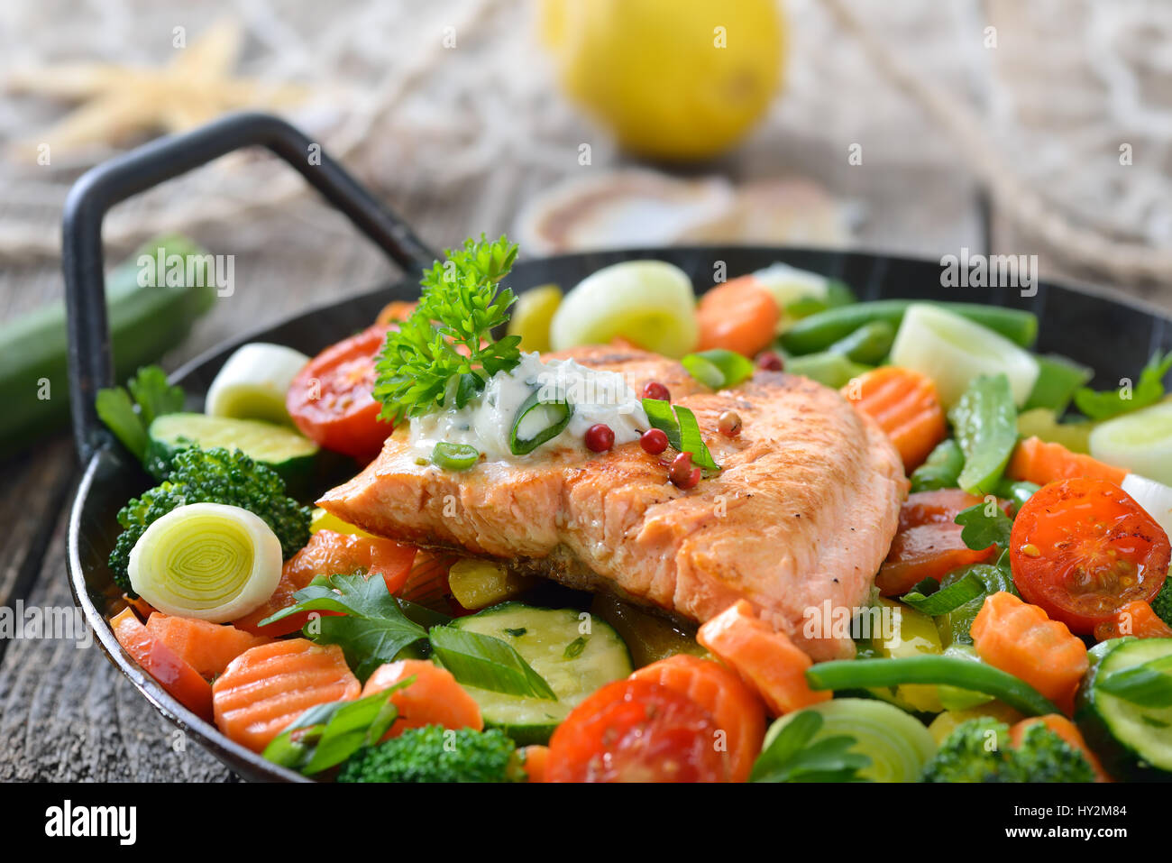 Fried salmon fillet on mixed colorful vegetables served in a frying pan,  a lemon and a fishing net with a starfish and seashells in the background Stock Photo