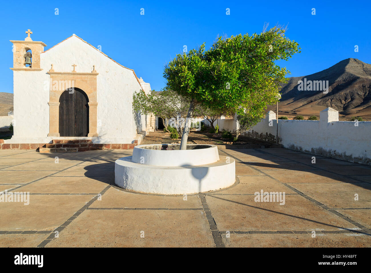 Well on courtyard of Typical white Canary style church in Tefia village and volcano mountain in background, Fuerteventura, Canary Islands, Spain Stock Photo