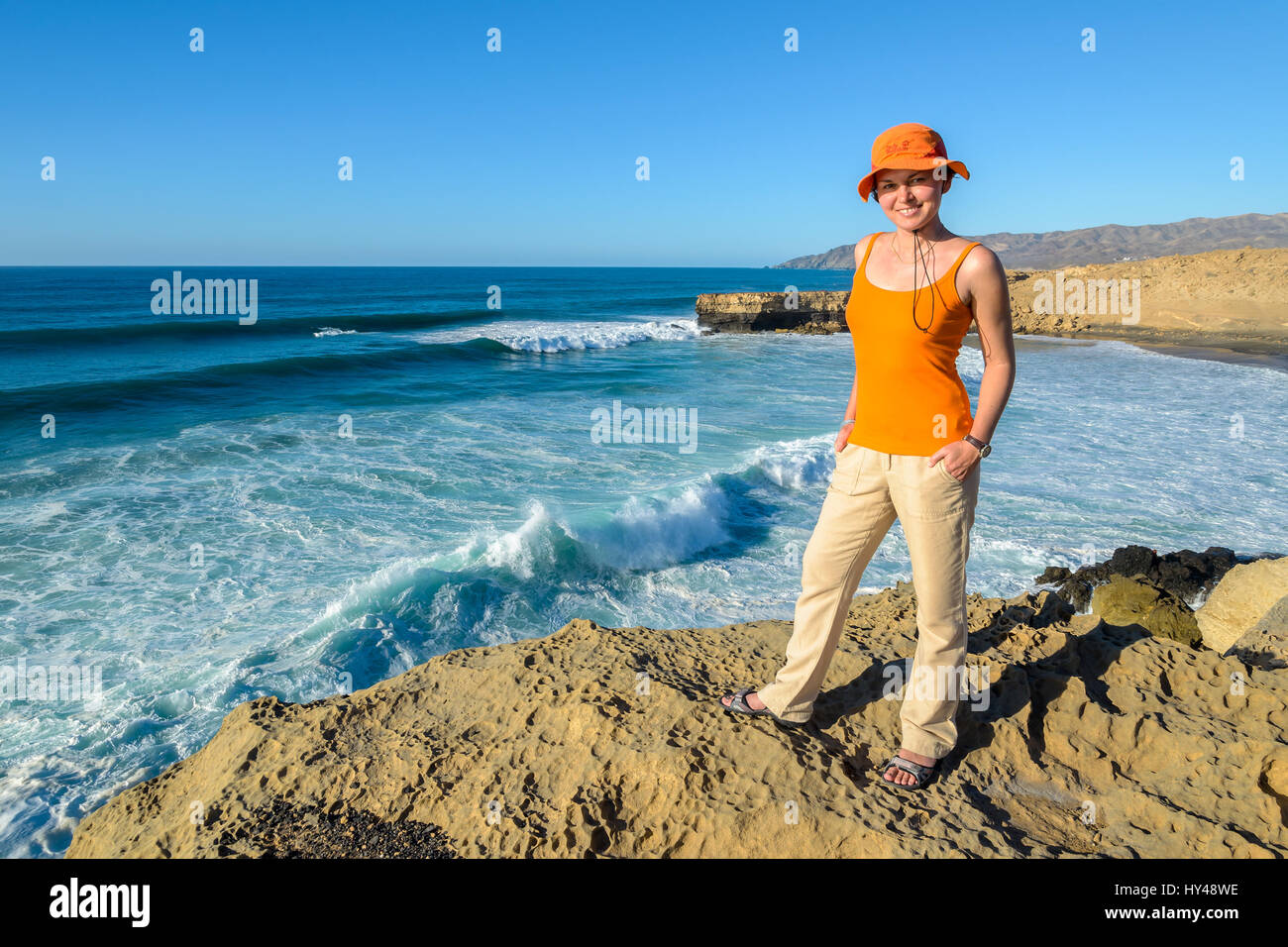 Young woman tourist standing on cliff and looking at camera near beautiful sea bay, Fuerteventura island, Spain Stock Photo