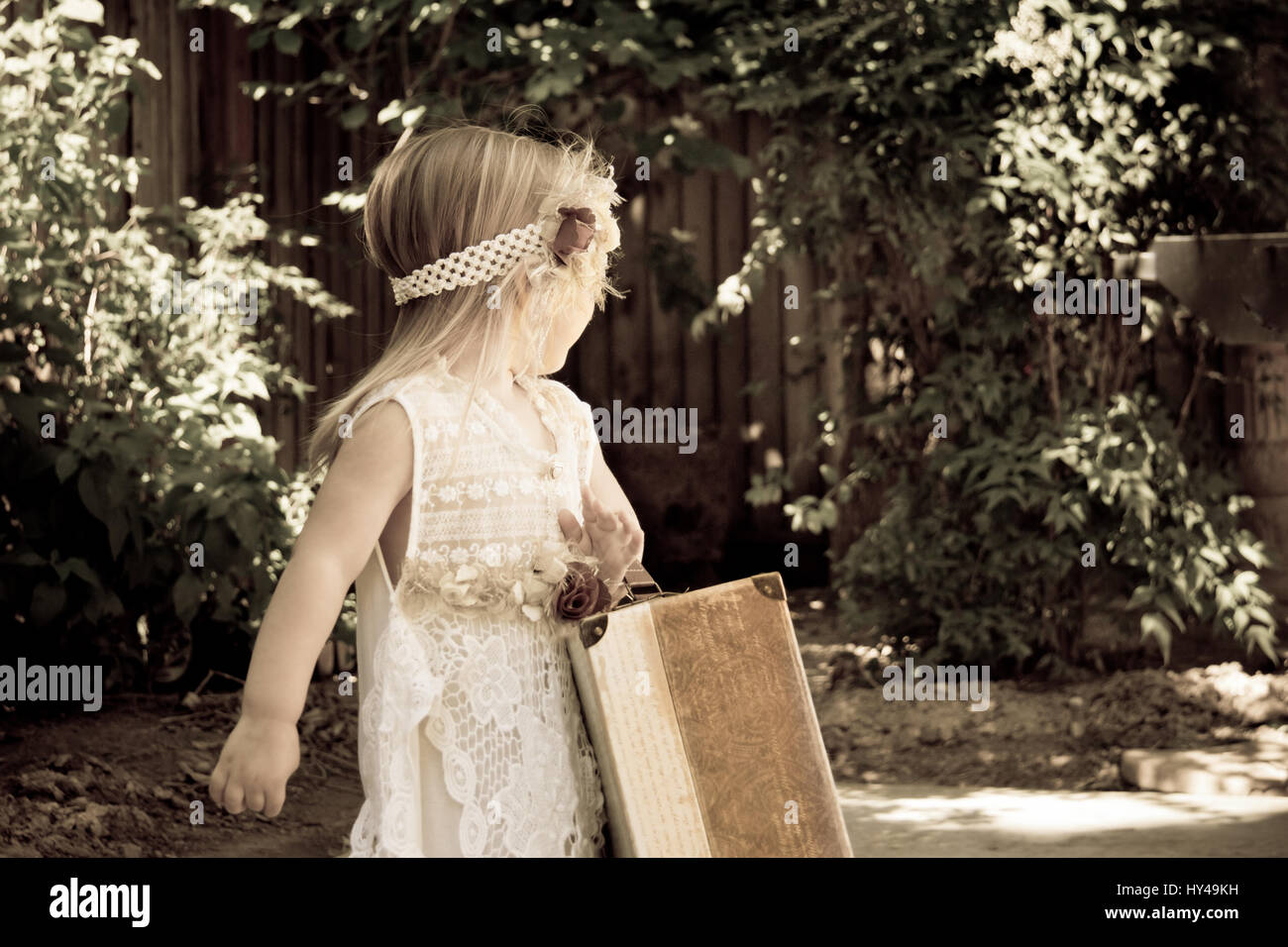 Little blonde toddler girl with peasant dress and luggage looking back over shoulder Stock Photo