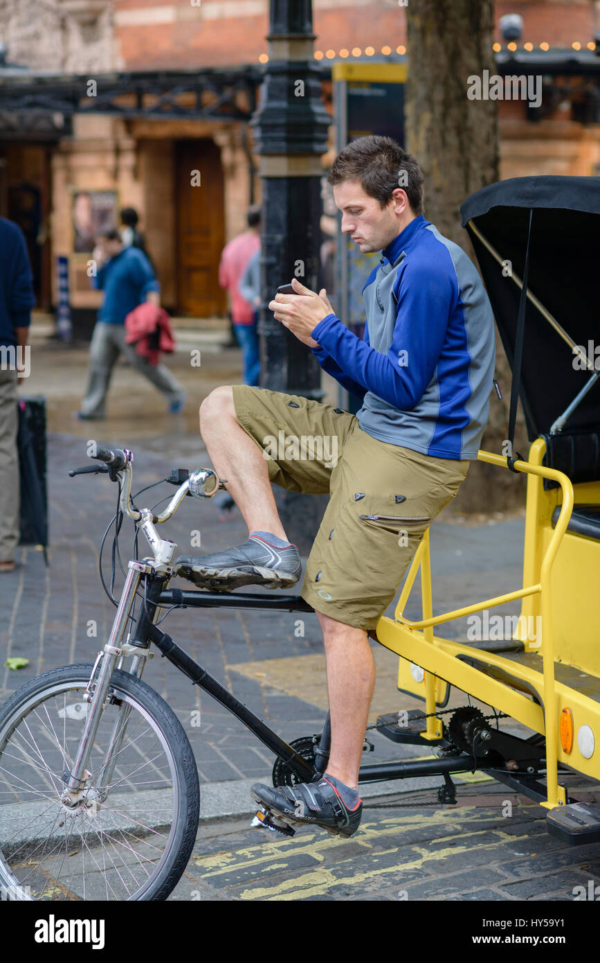 Young male London pedicab driver (rickshaw rider) checks his mobile phone while waiting for his next fare. Young man using smartphone at work, wait Stock Photo