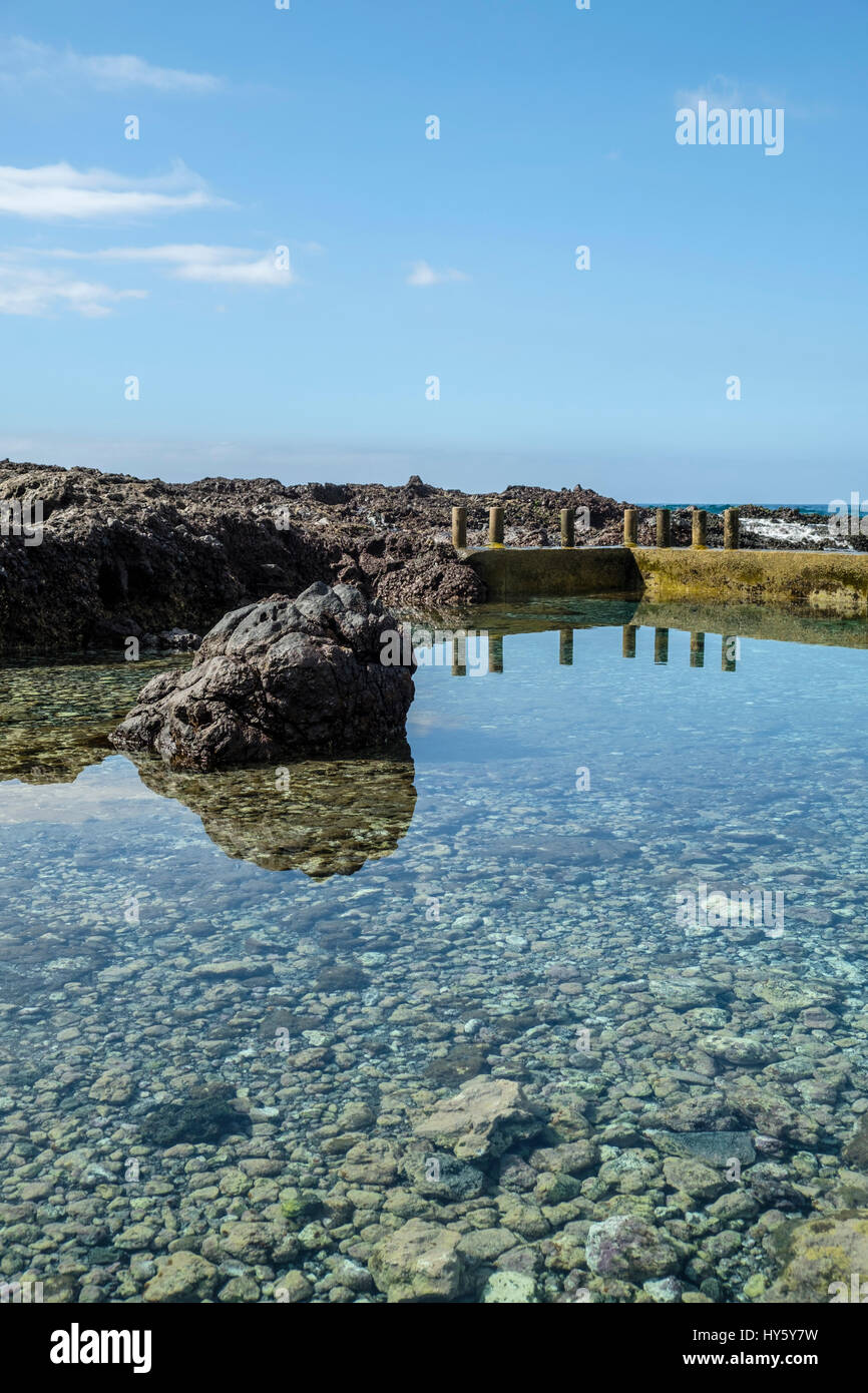 Still clear blue waters of a natural rock pool on the Tenerife coast near Alcala with reflections of a concrete sea wall and large black volcanic rock Stock Photo
