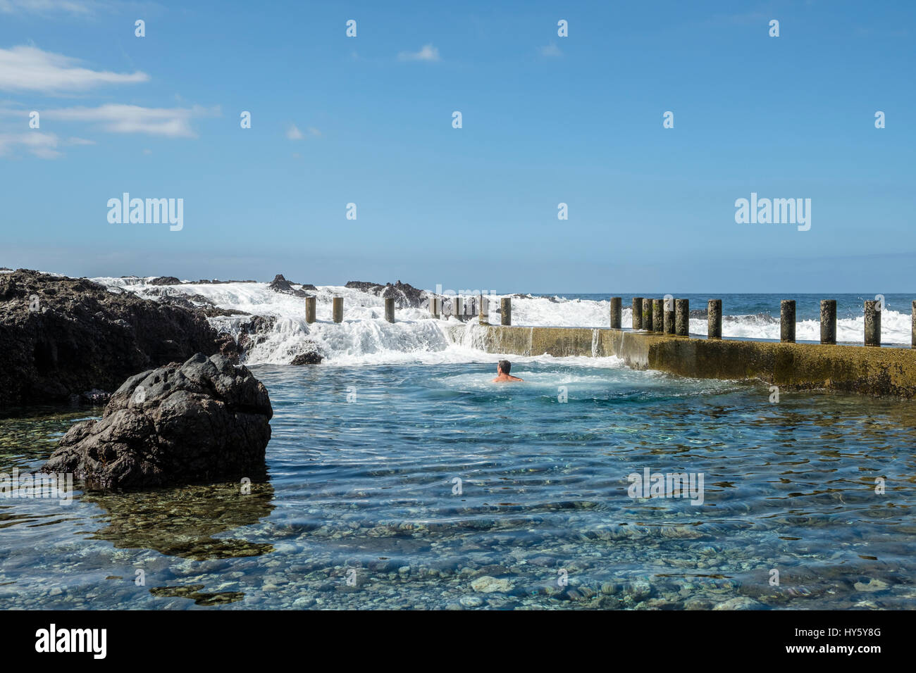 A swimmer enjoys a refreshing dip in the still clear blue waters of a natural rock pool on the Tenerife coast near Alcala while waves from the Atlanti Stock Photo