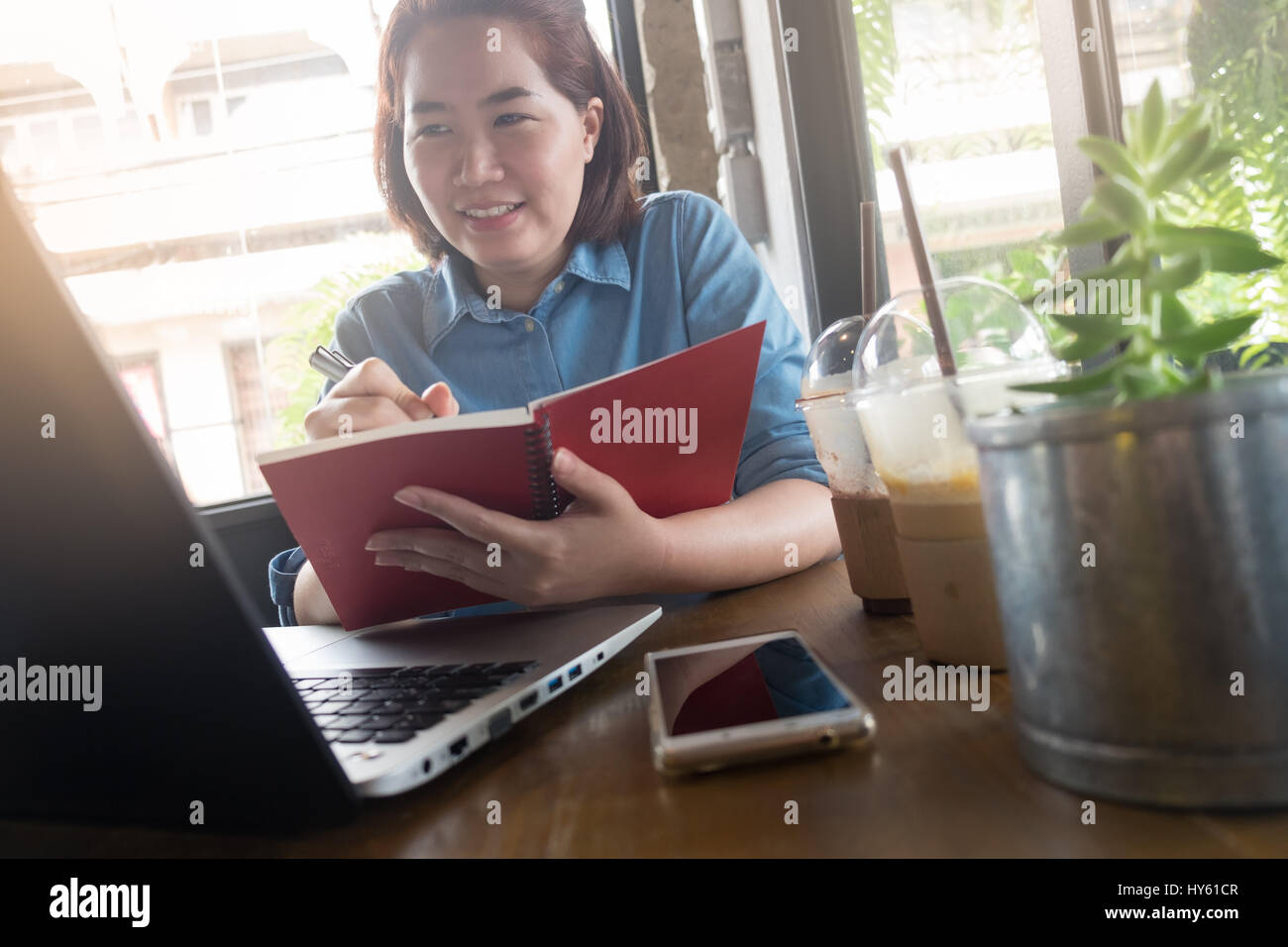 Young Asian hipster woman writing on notebook while working with laptop computer in coffee shop. Freelance business starup activity concept Stock Photo
