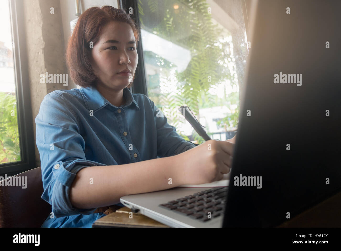 Young Asian hipster woman writing on notebook while working with laptop computer in coffee shop. Freelance business starup activity concept Stock Photo