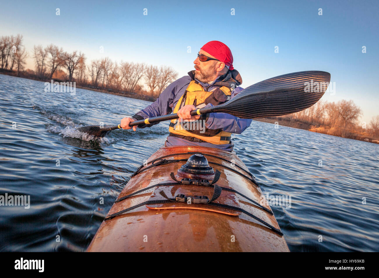 mature male paddler exercising (turning boat using rudder stroke with his wing carbon fiber paddle) in a home built wooden sea kayak on lake, fall sce Stock Photo
