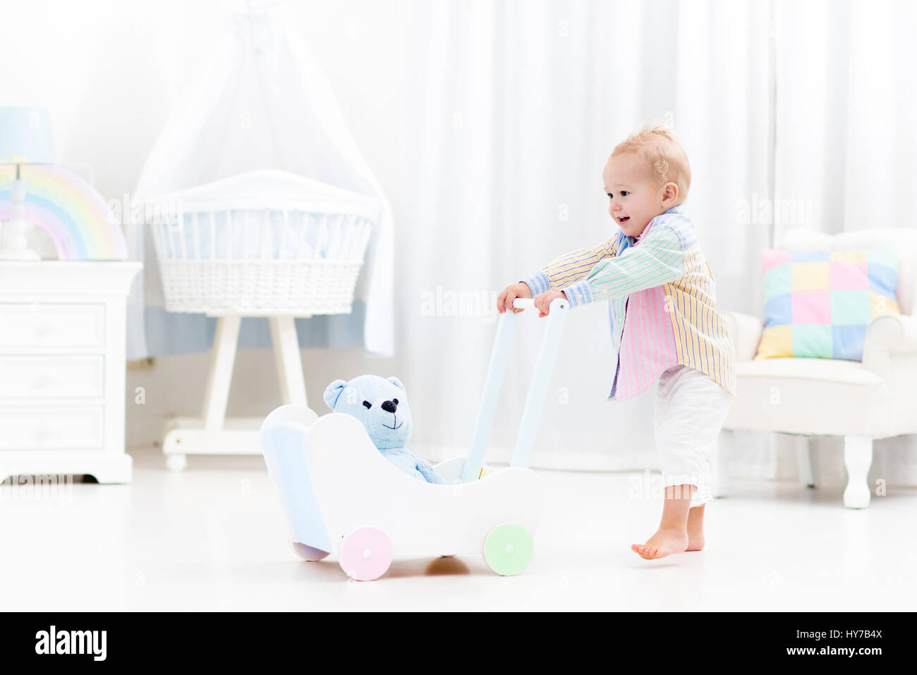 Baby boy learning to walk with wooden push walker in white bedroom with
