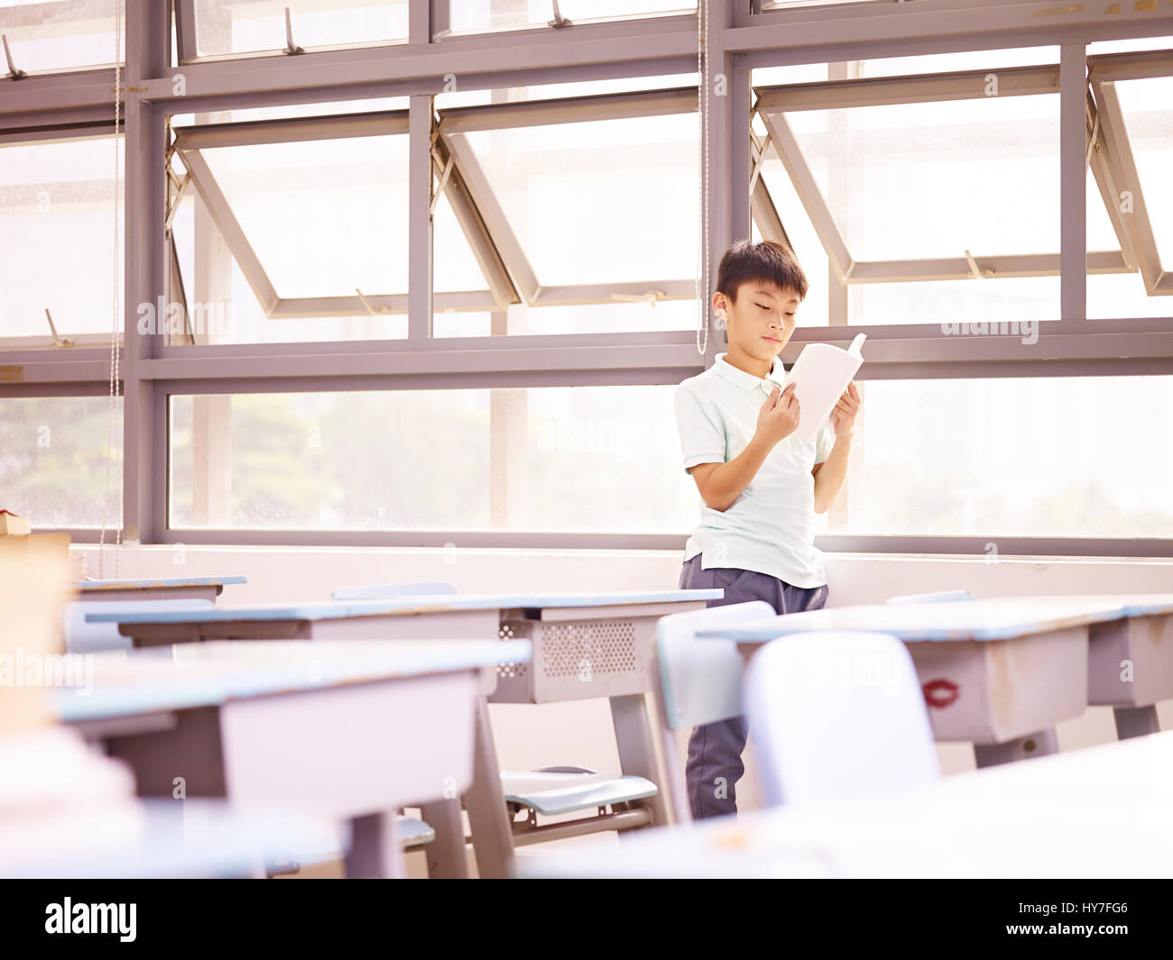 asian elementary schoolchild standing by the windows reading a book in classroom. Stock Photo