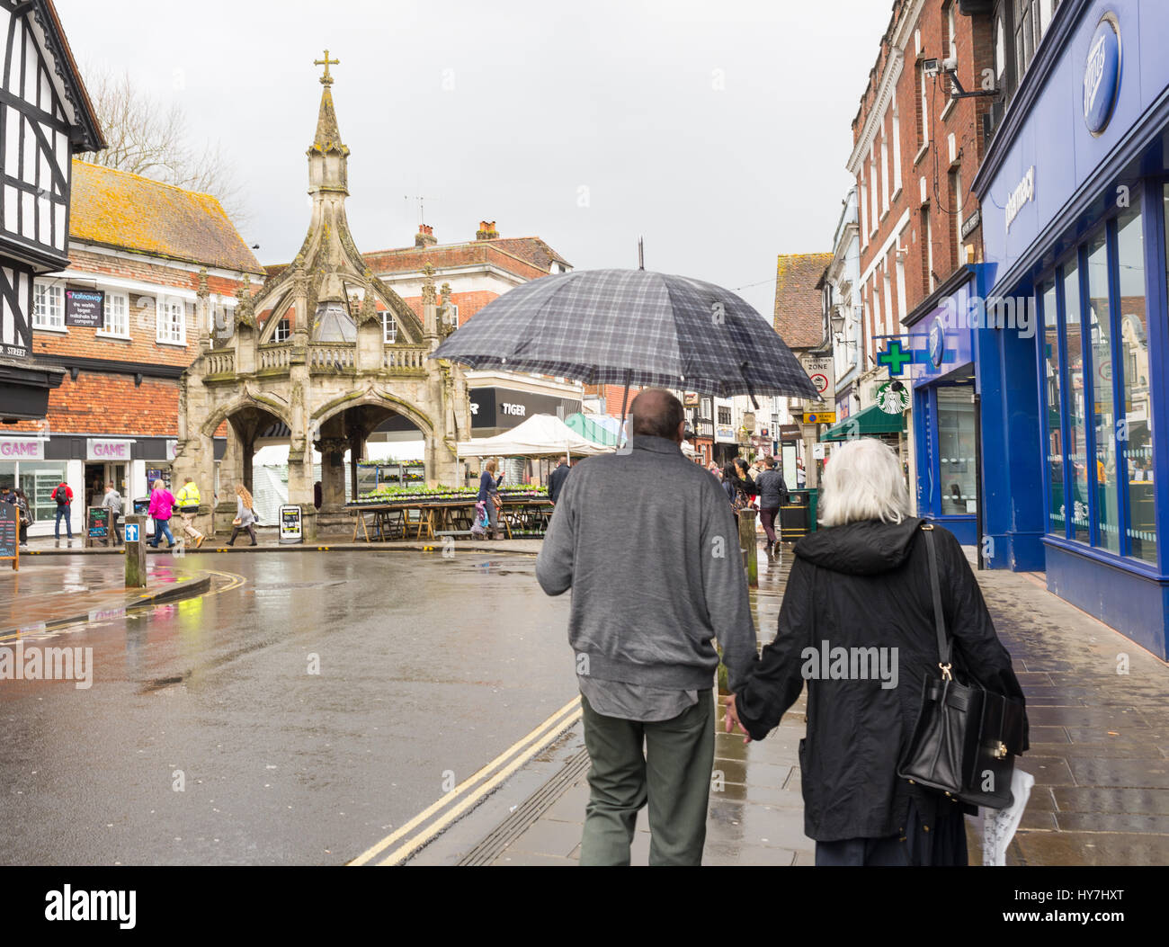Elderly couple using an umbrella on Blue Boar Row near the Poultry Cross ancient monument, Salisbury, Wiltshire, UK. Mixed weather in the historic cathedral city with heavy April rain showers and sunshine on a spring afternoon. Stock Photo