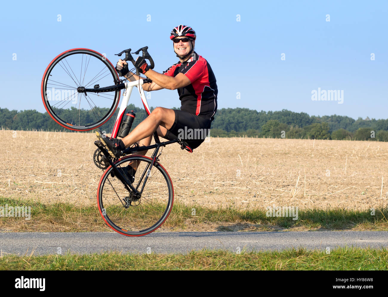 Cheerful cyclist riding on the rear wheel. Biker balancing while driving on a road bike. Risky ride on one wheel. Stock Photo
