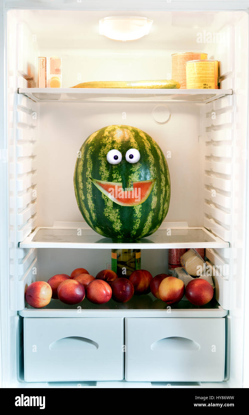 Watermelon on a shelf in the refrigerator. Cheerful funny watermelon with carved a smile looks out of fridge. Stock Photo