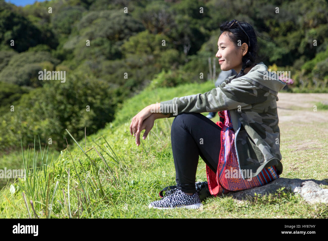 Young woman sitting on green grass. Asian girl resting on the lookout, watching the landscape. Tourist relaxes on a trip. Stock Photo