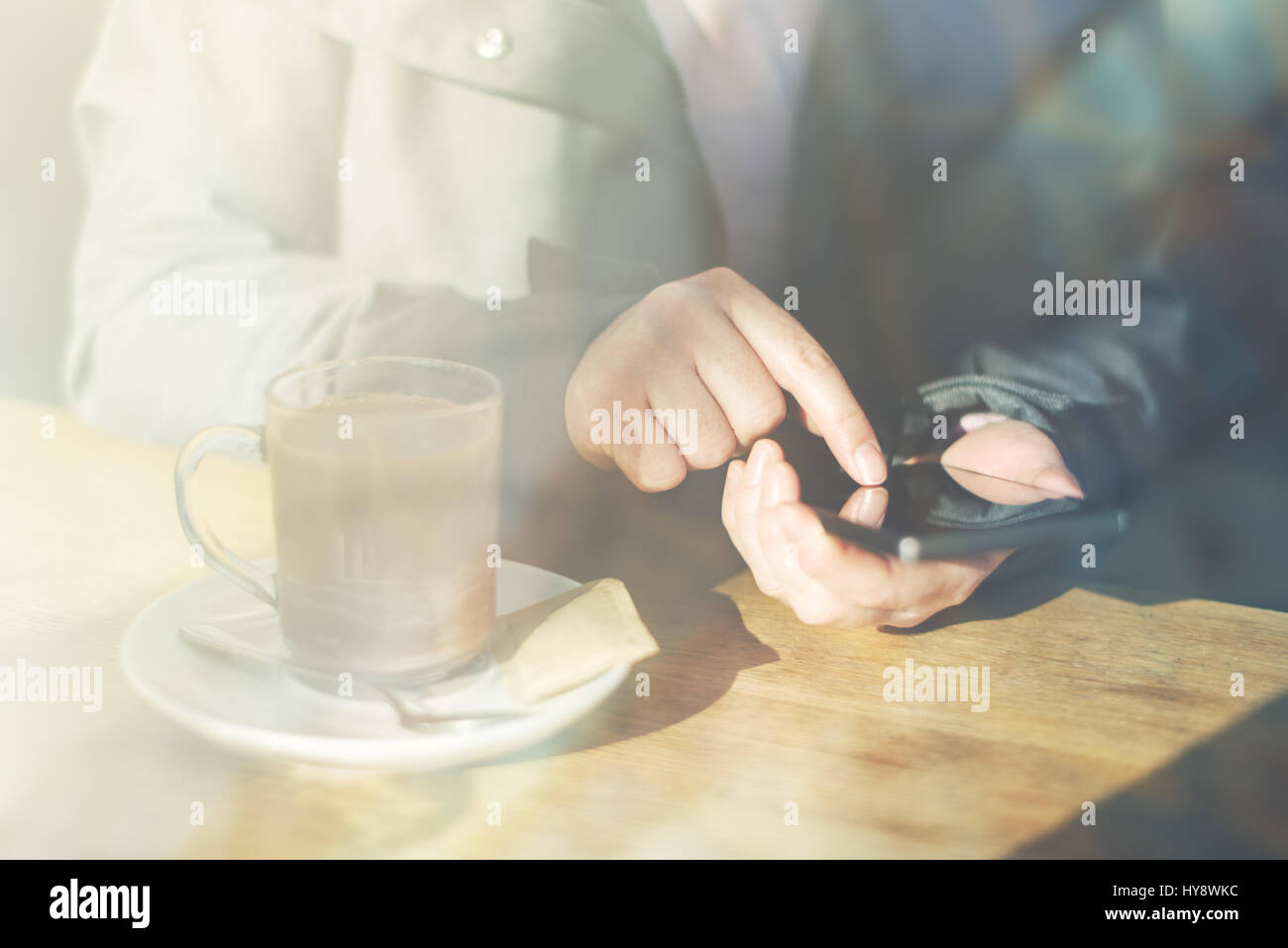 Woman using mobile phone and drinking hot chocolate in cafe. Casual young adult caucasian female behind the glass typing text message on smartphone ap Stock Photo