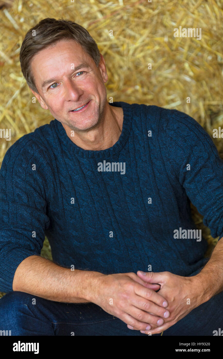 Portrait shot of an attractive, successful and happy middle aged man male wearing a blue sweater sitting on hay bales in a barn or stables Stock Photo