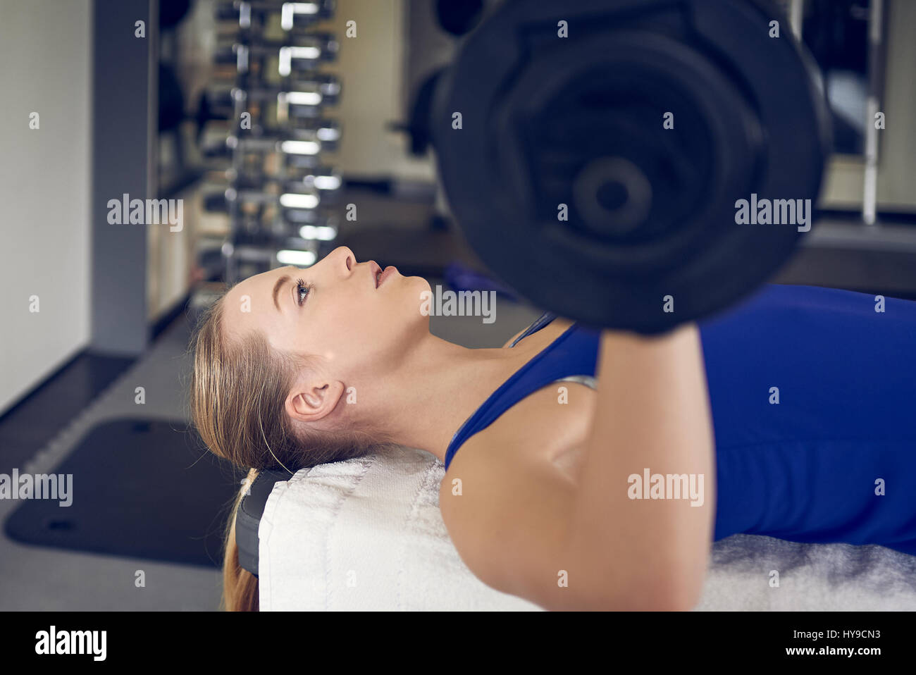 Close-up side view of young blonde woman in blue top doing bench pressing exercise with bar-bell in gym Stock Photo