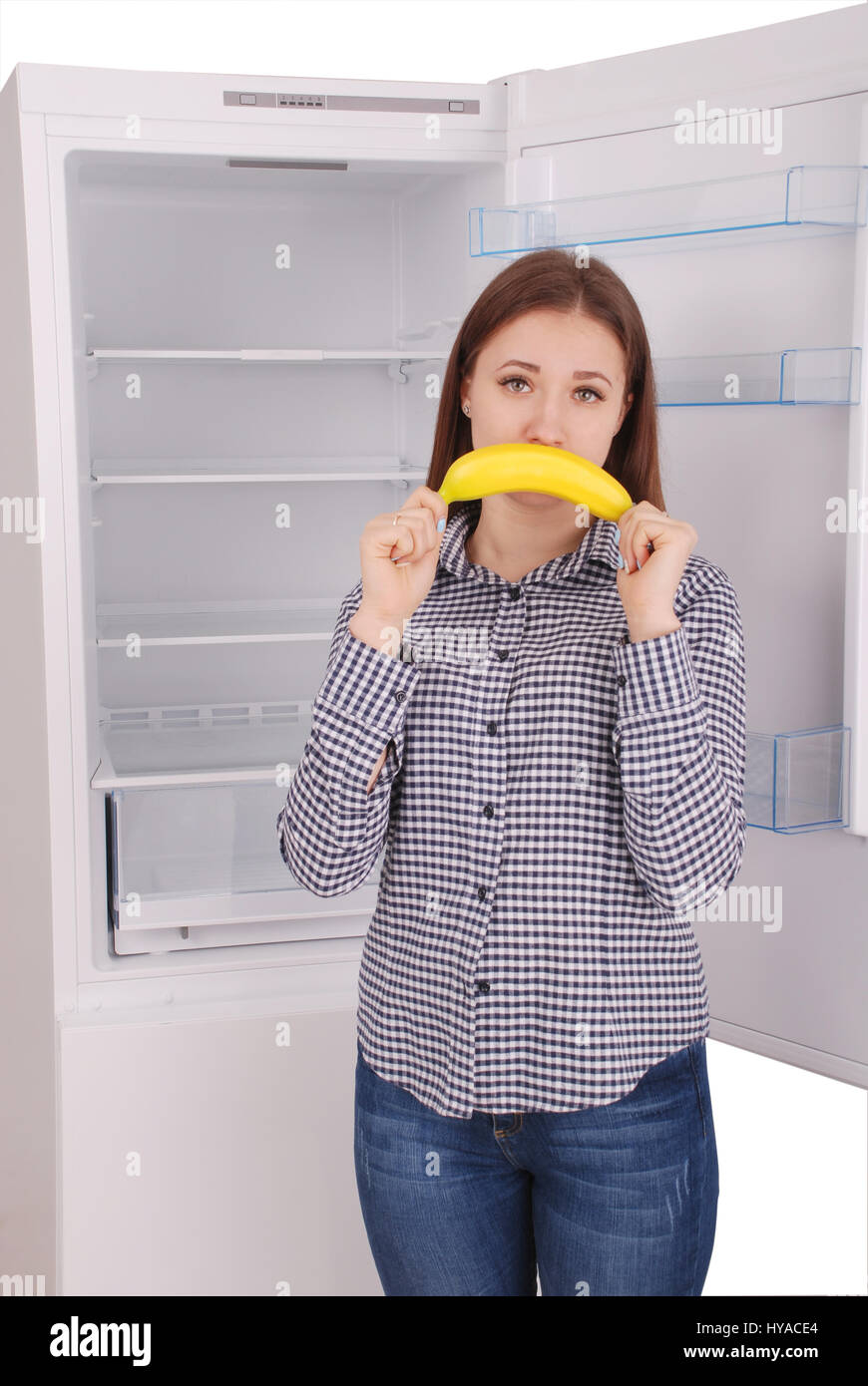 A young girl holds up a banana to her mouth, imitating a smile. Beautiful young girl near the fridge. Stock Photo