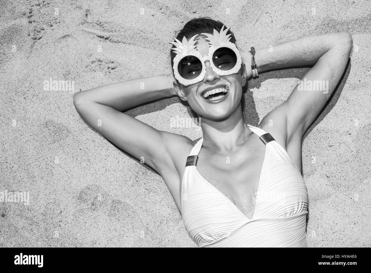 Warm sand treatment. smiling woman in swimsuit and funny pineapple glasses laying on the sand Stock Photo