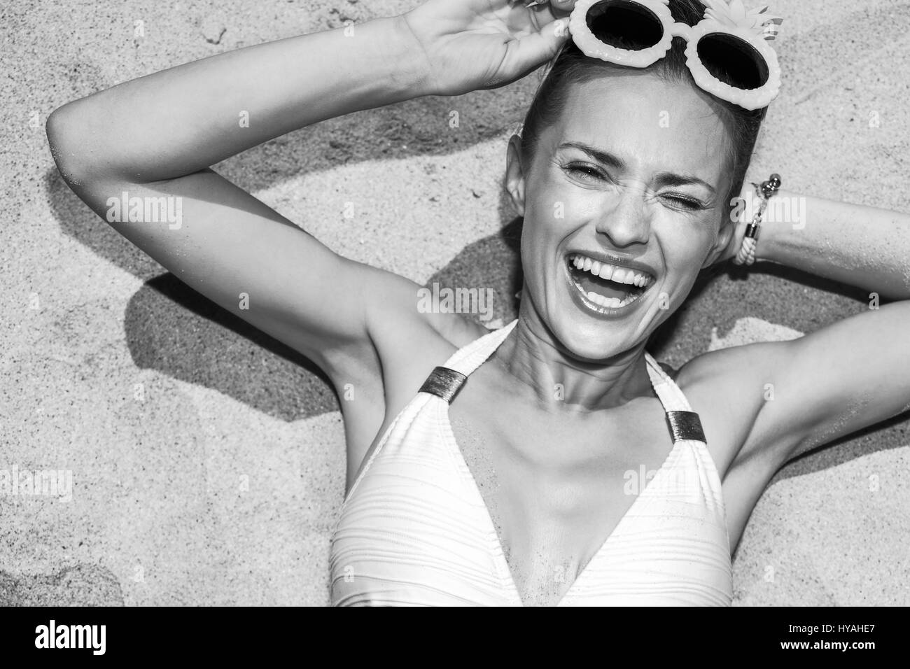 Warm sand treatment. Portrait of smiling woman in swimsuit with funny pineapple glasses laying on the sand and winking Stock Photo