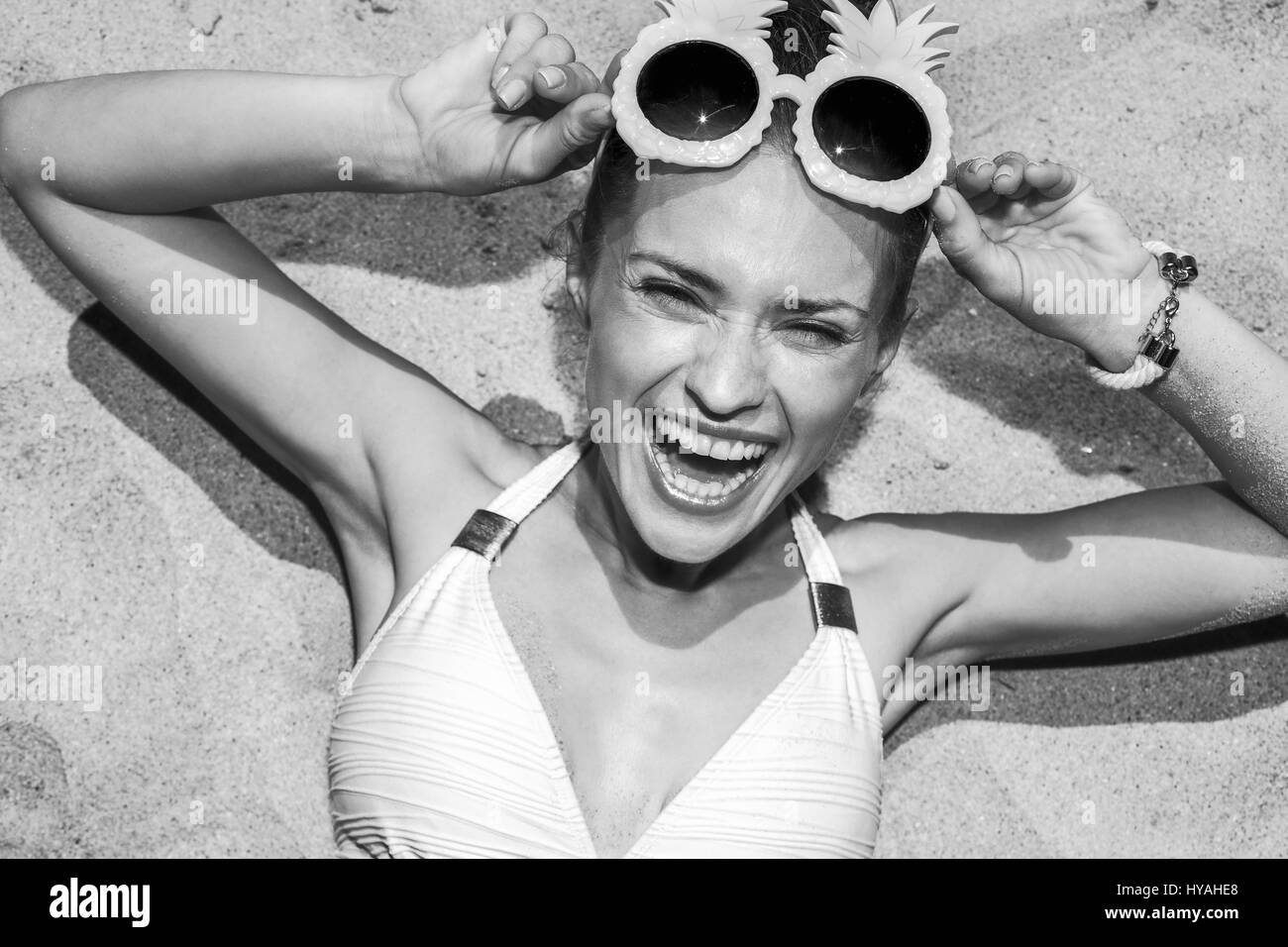 Warm sand treatment. Portrait of cheerful woman in swimsuit with funny pineapple glasses laying on the sand Stock Photo