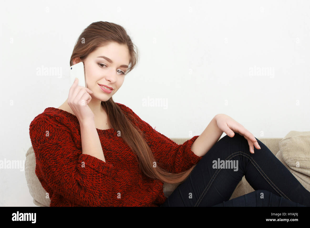 Young beautiful cheerful red-haired girl in a blue jeans jacket sitting on the couch at home and talking on a cell phone. Stock Photo