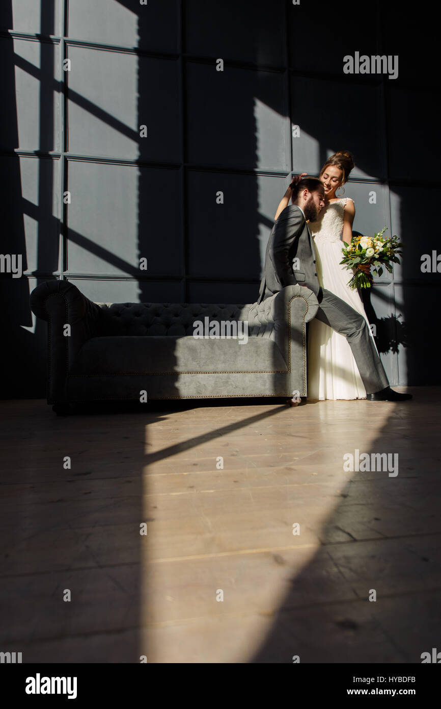 The bride smiling embraces the groom sitting on the arm of the sofa ...