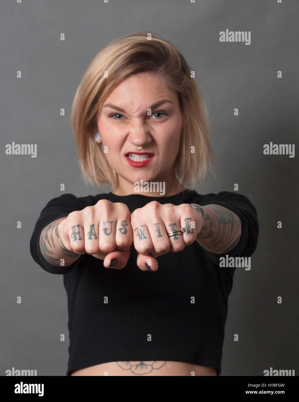 Young woman with angry facial expression, holding her hands in front of her, the hands with Hause wife tatooe on her fingers. Wearing black t-shirt, r Stock Photo