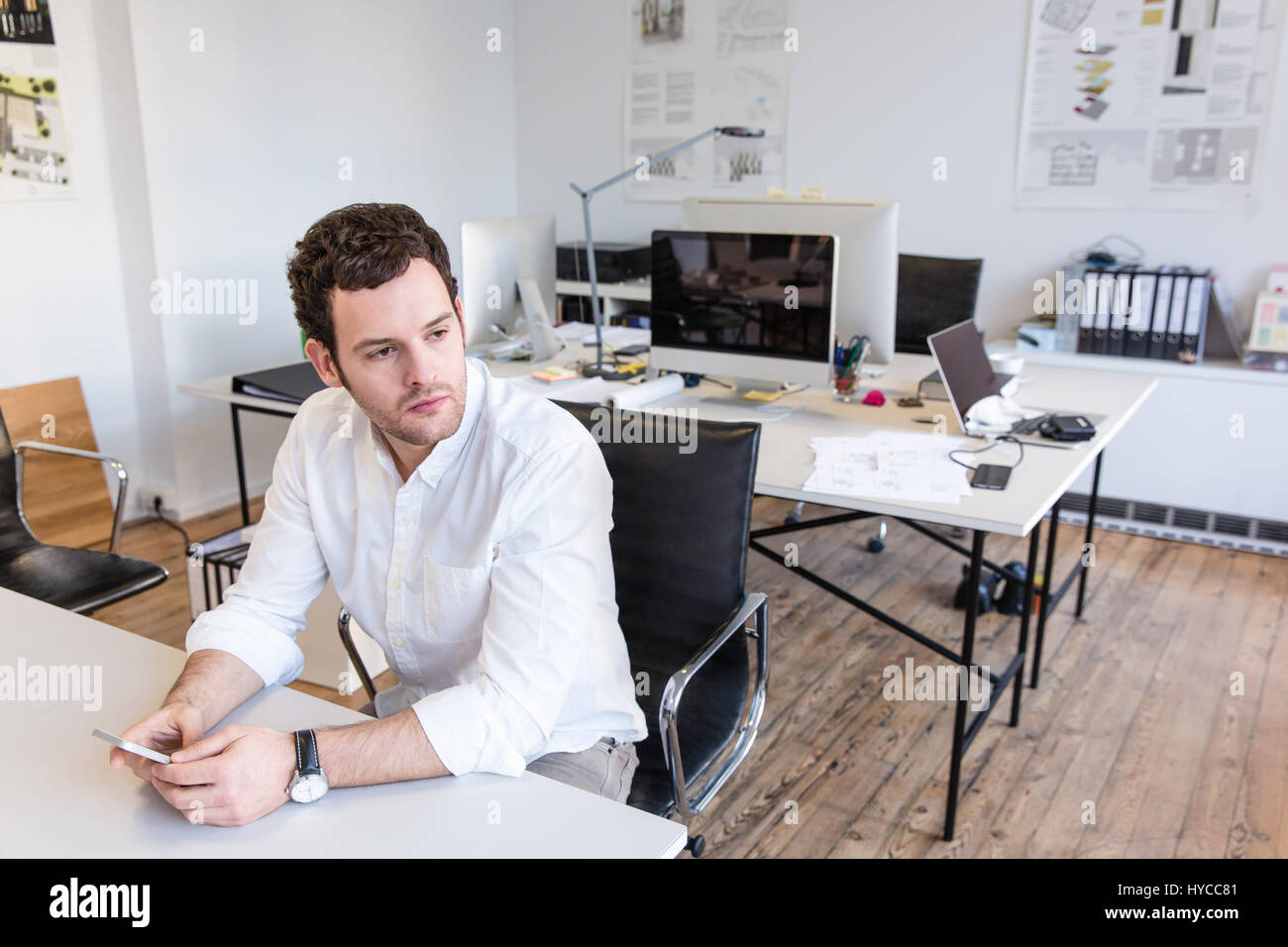 Mid adult man sitting in office at desk holding smartphone looking away Stock Photo