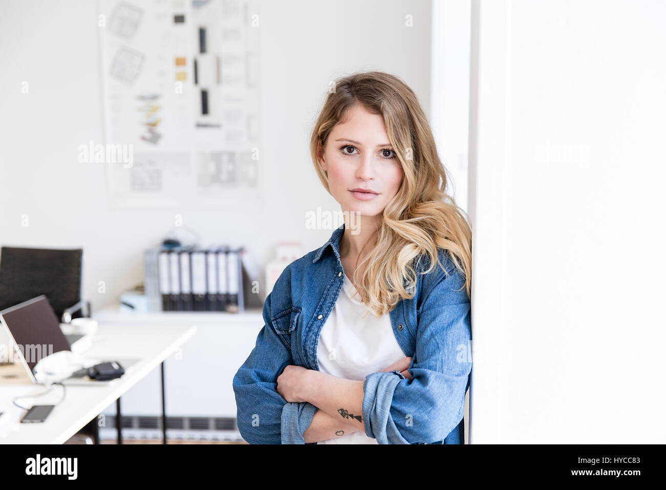 Young woman in office leaning against wall arms crossed looking at camera Stock Photo