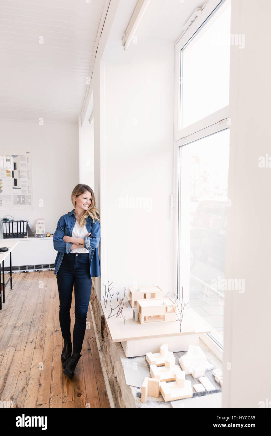 Full length view of young woman in office leaning against wall arms crossed looking away out of window Stock Photo