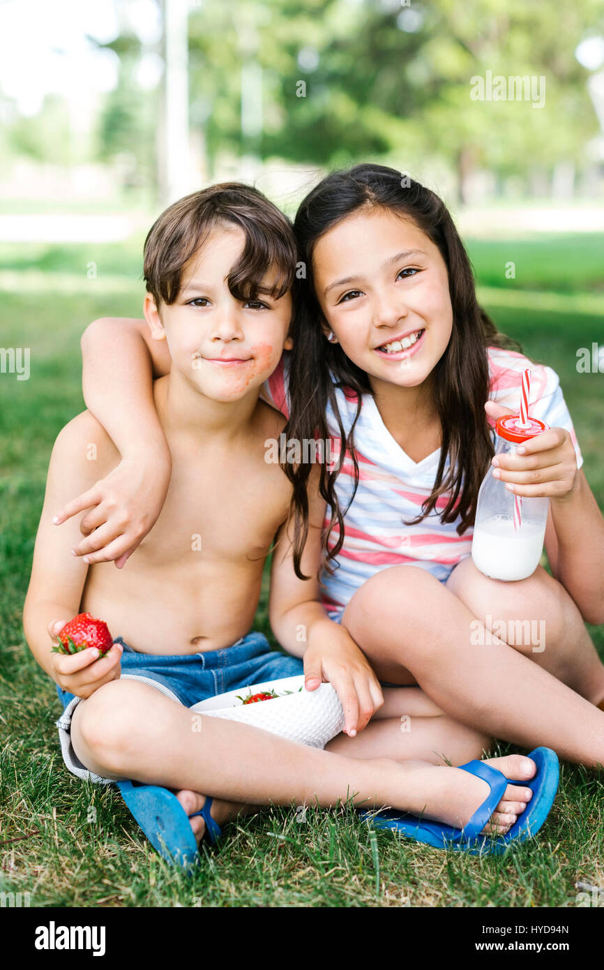 Boy (6-7) and girl (10-11) sitting arm around on grass in summer Stock Photo