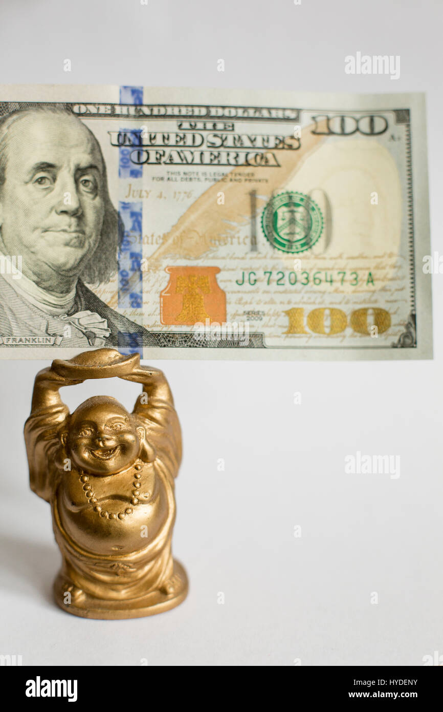 a small golden buddha figurine standing on a white back ground holds a one hundred dollar bill in United States currency above his head Stock Photo