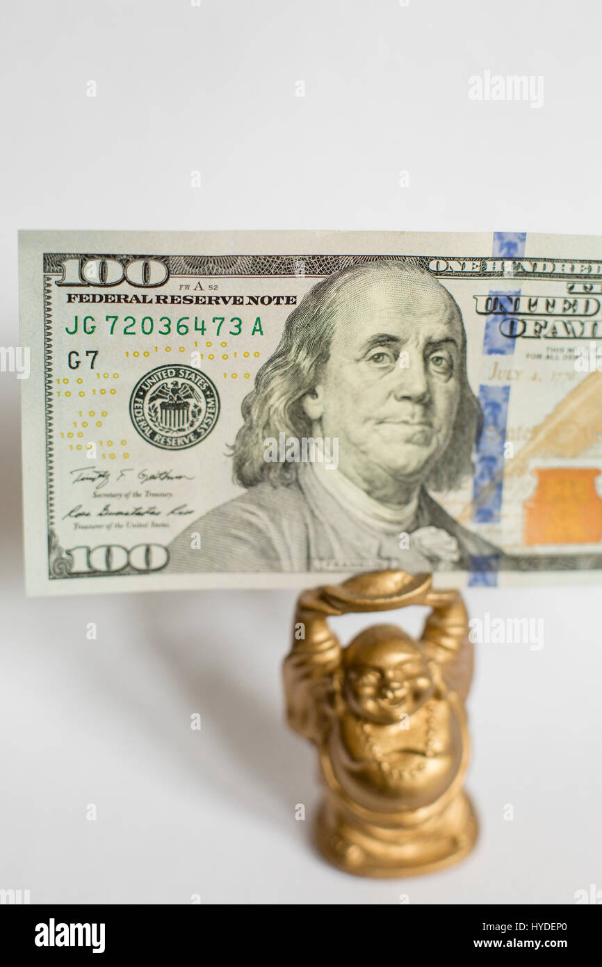 a small golden buddha figurine standing on a white back ground holds a one hundred dollar bill in United States currency above his head Stock Photo